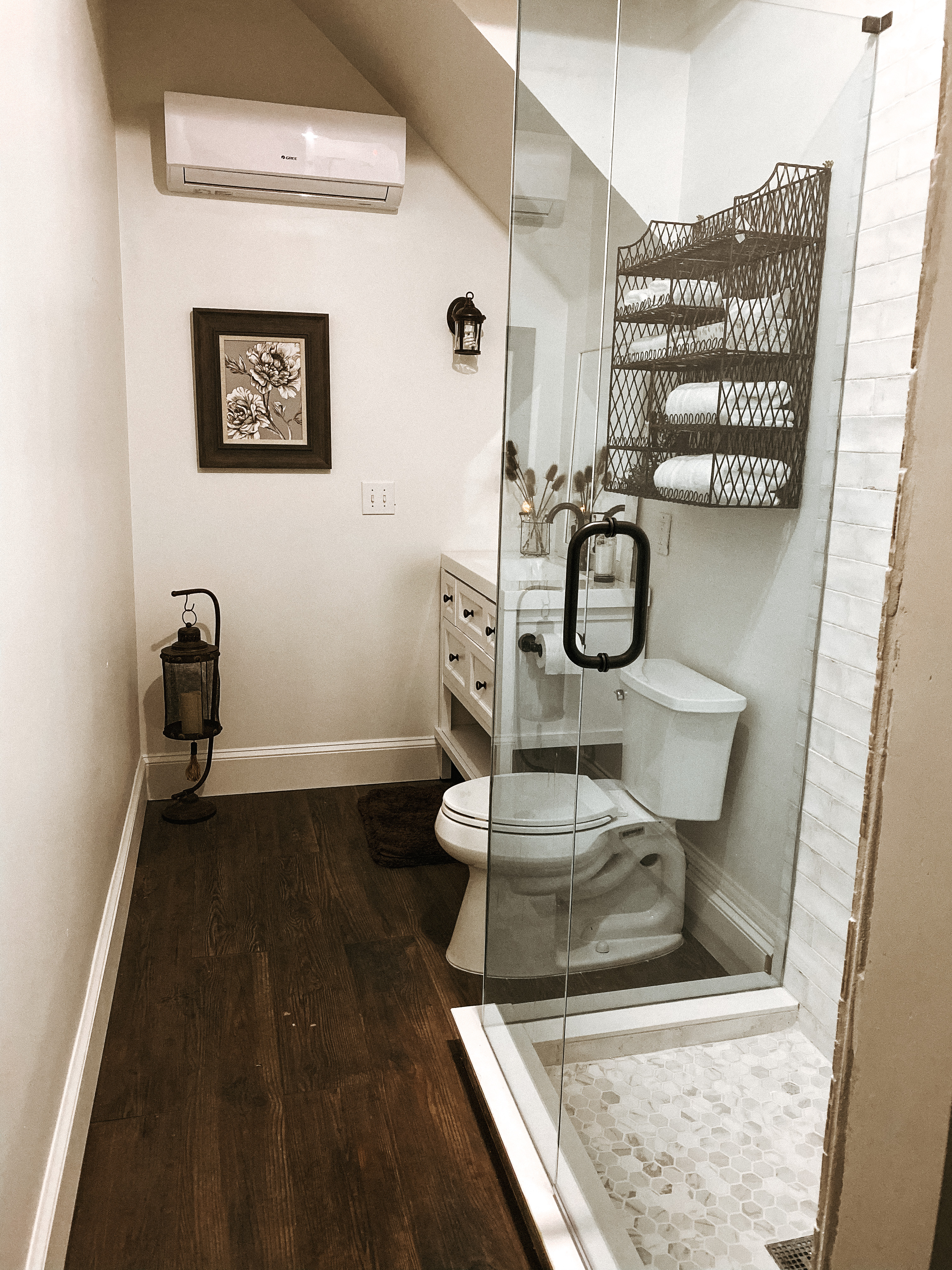 A compact, modern bathroom featuring dark wood floors, a glass-enclosed walk-in shower with pebble tiling, a white vanity, and a wall-mounted towel rack beneath a sloped ceiling.