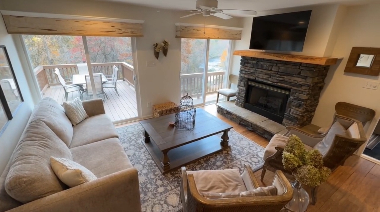 An airy living room featuring a large stone fireplace with a wooden mantel, comfortable tan seating, and sliding glass doors that lead to an outdoor deck with a dining set.