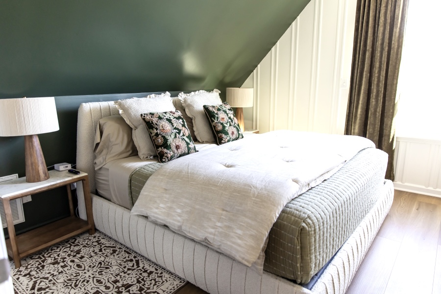 A cozy bedroom featuring a plush white bed with floral-patterned pillows against a deep green accent wall. The room is set in an attic with a sloped ceiling and includes rustic wooden nightstands and a large window with gold curtains.