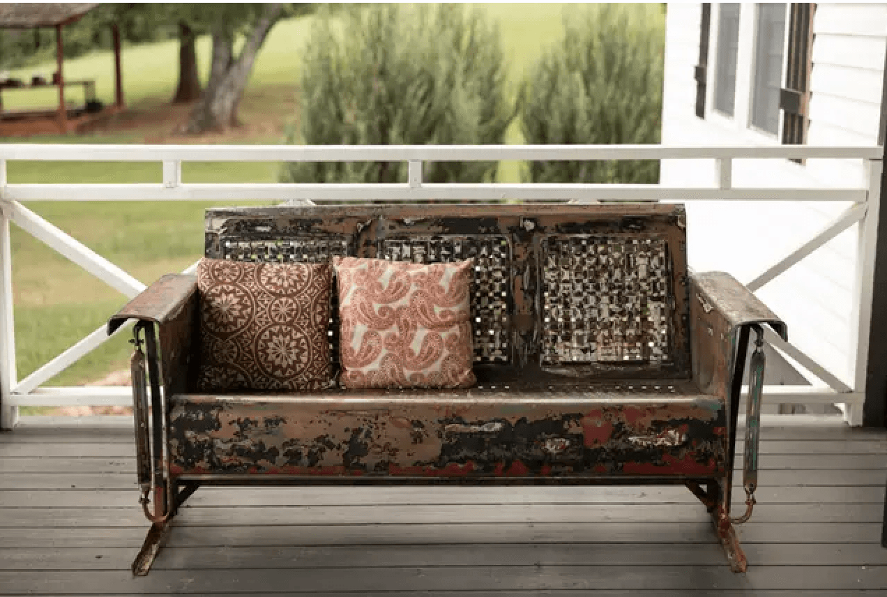 A weathered porch swing with decorative pillows, set against a green outdoor backdrop.