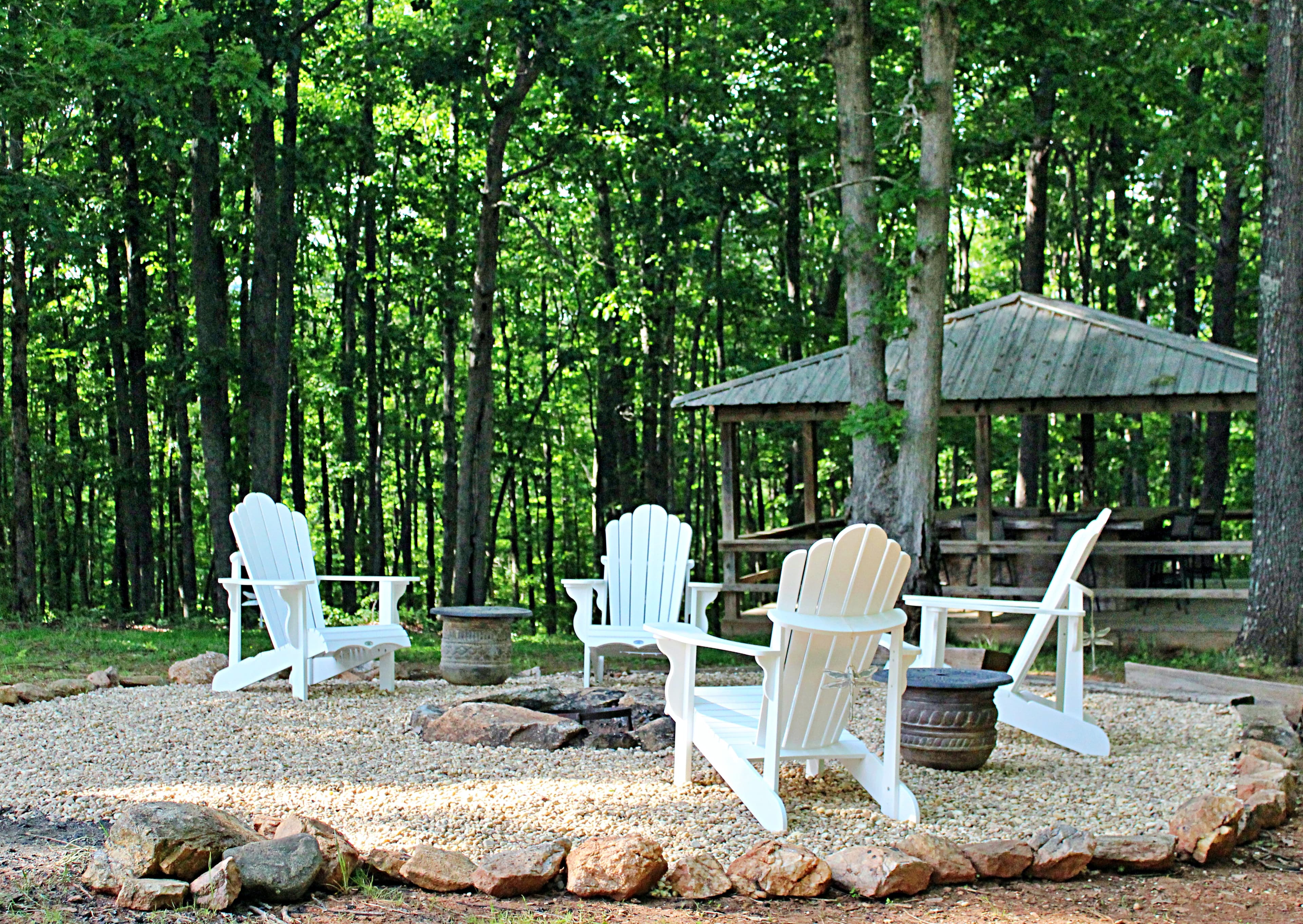 Four white Adirondack chairs encircle a stone fire pit on a gravel terrace, with a rustic wooden pavilion and a dense green forest in the background.
