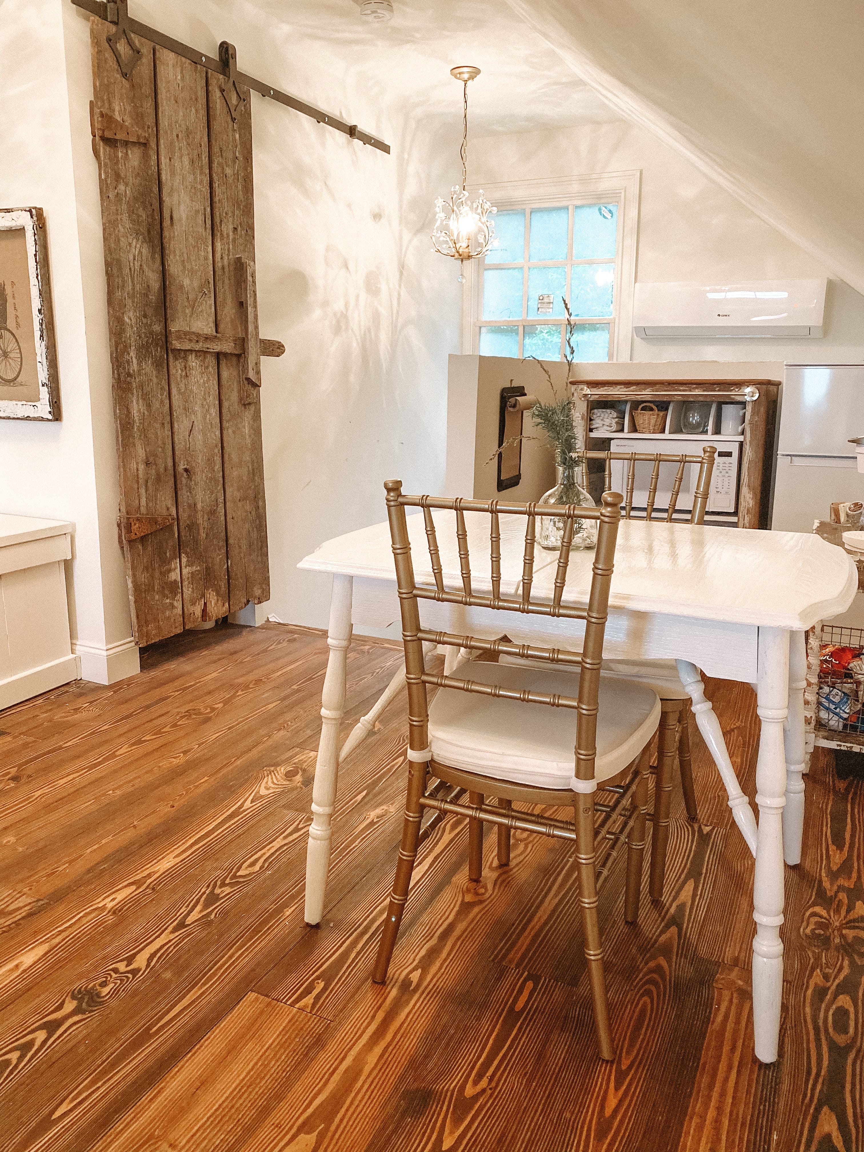 A white dining table with two gold-colored chairs sits on rustic hardwood floors, positioned in front of a kitchenette and a weathered wood barn-style sliding door.