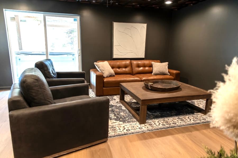 A modern living room featuring charcoal-grey walls, a brown leather sofa, and two matching grey armchairs centered around a large wooden coffee table. The space includes a patterned blue and white area rug and a sliding glass door leading outside.