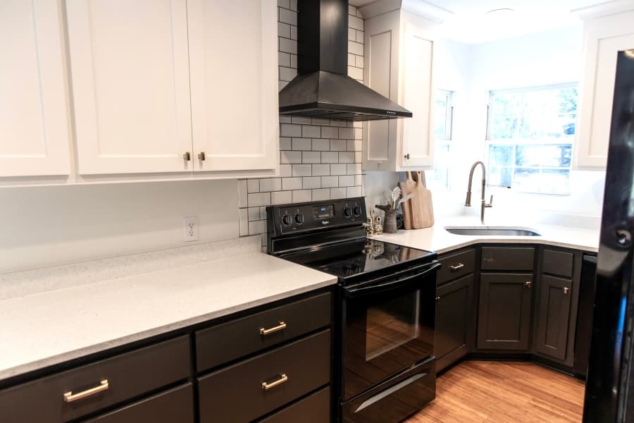 A modern kitchen corner featuring white upper cabinets, dark lower cabinetry with gold handles, and a white subway tile backsplash. The space includes a black electric stove with a matching range hood and a corner sink positioned in front of a bright window.