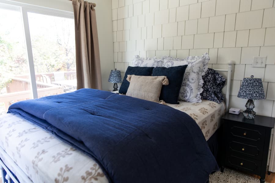 A cozy bedroom featuring a navy blue comforter on a white bed frame, set against a cream-colored shingle-style accent wall. The room includes dark nightstands with matching patterned lamps and a large sliding glass door that overlooks an outdoor deck.