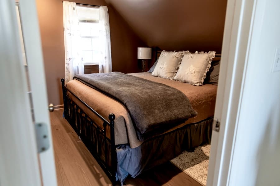 A cozy bedroom seen through a doorway, featuring a black metal bed frame with brown and charcoal bedding. The room has dark brown walls, a sloped ceiling, and a bright window with white lace curtains.