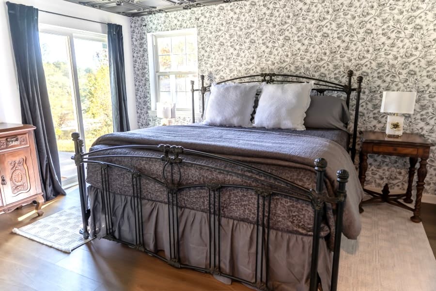 An elegant bedroom featuring a dark metal bed frame with grey patterned bedding, set against a floral wallpaper accent wall. The room includes a vintage wooden nightstand and a sliding glass door with dark curtains leading to a sunlit outdoor area.