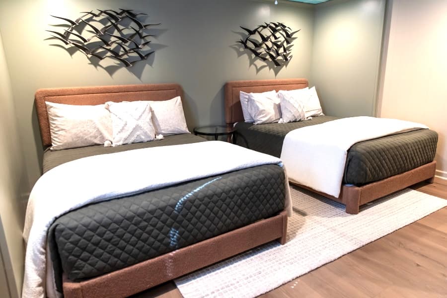Two identical queen-sized beds with grey quilted mattresses and white pillows set against a light green wall featuring metal bird-shaped wall art.