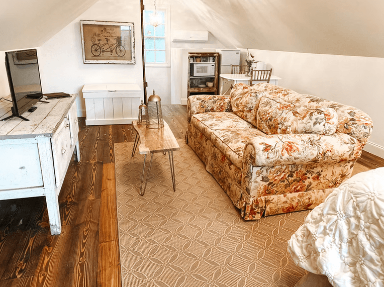 A cozy attic living space featuring a floral-patterned sofa, rustic hardwood floors, and a white media console with a television beneath a sloped ceiling.