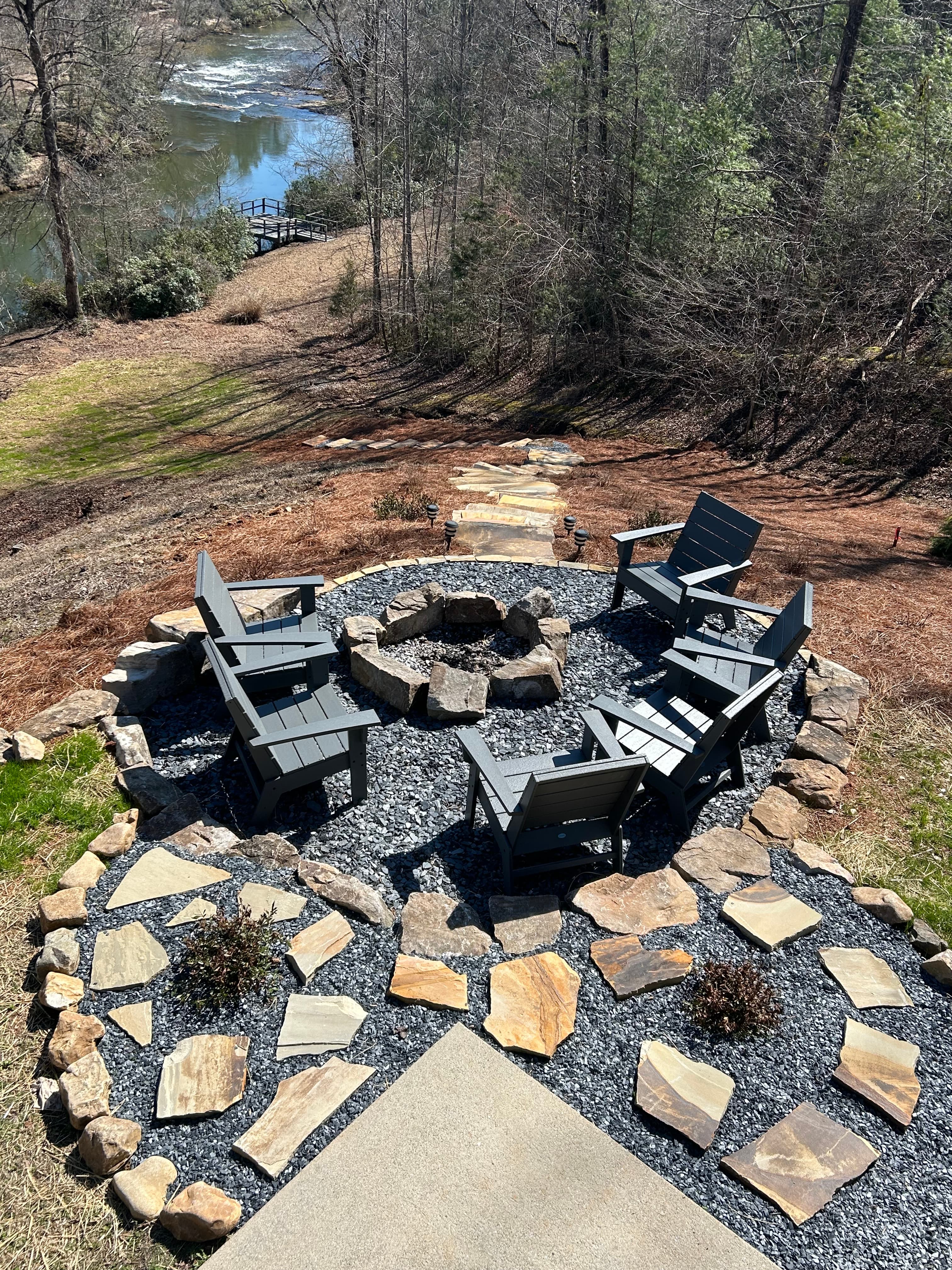 Outdoor firepit with 6 gray chairs that overlooks a river.