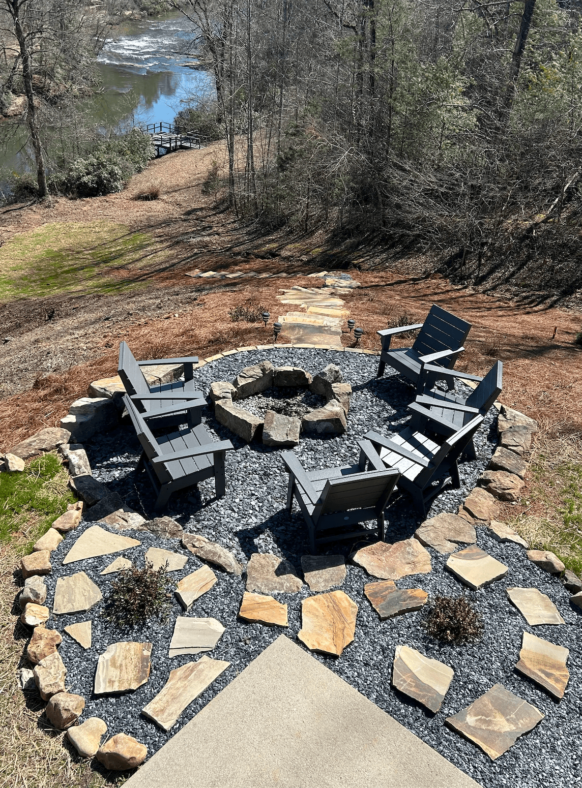 A fire pit area surrounded by stone seating and a gravel base, overlooking a river and wooded landscape.