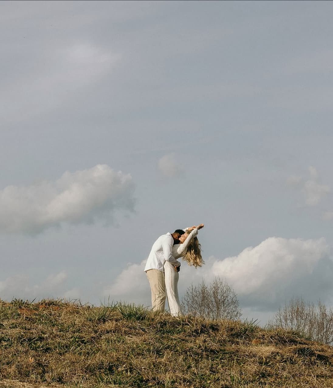 A couple embraces and kisses on a grassy hill against a cloudy sky.