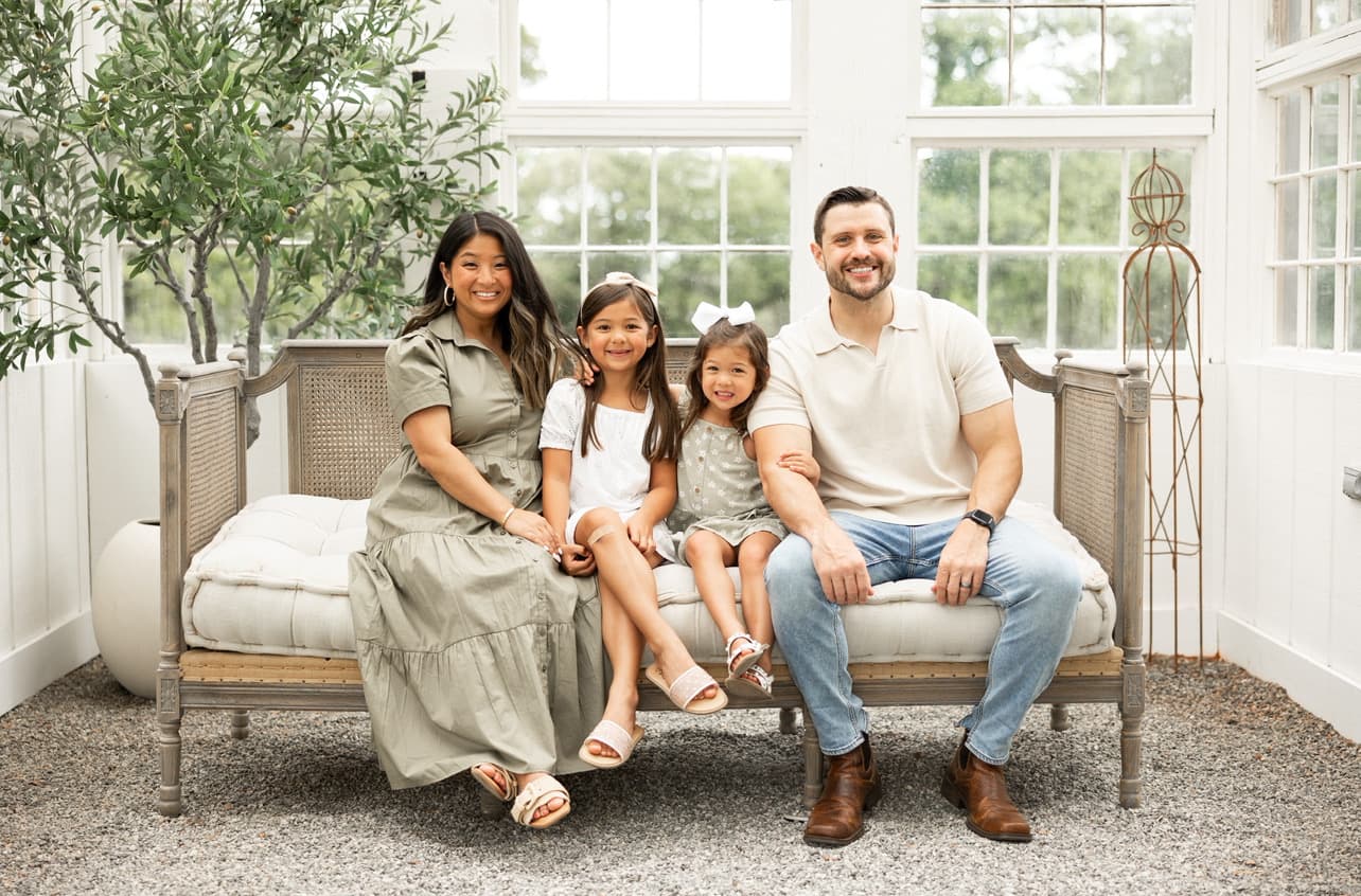 A family of four sits together on a couch in a bright, airy space filled with greenery.