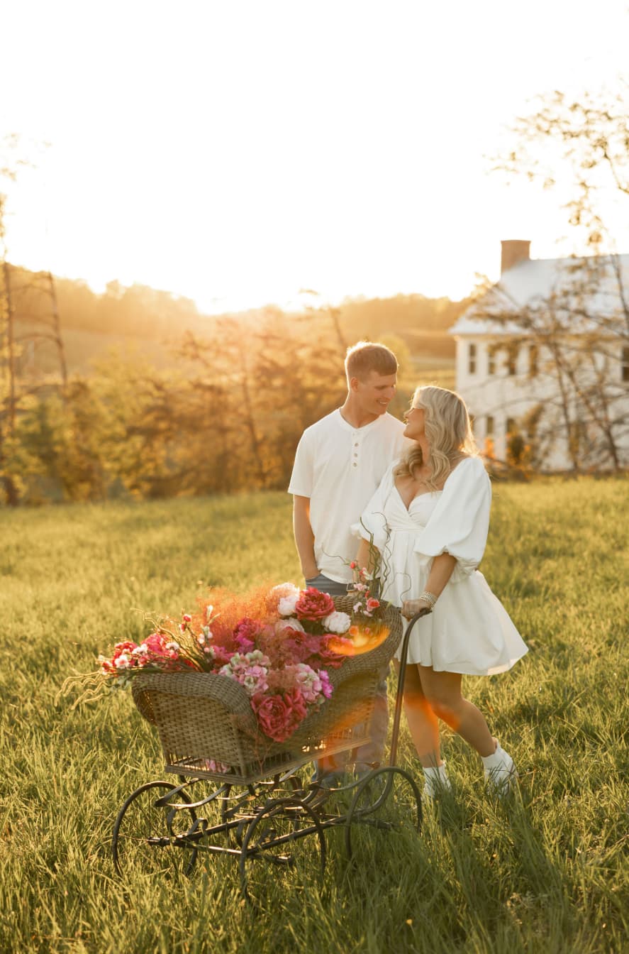 A couple stands together in a sunlit field, with a cart filled with colorful flowers.