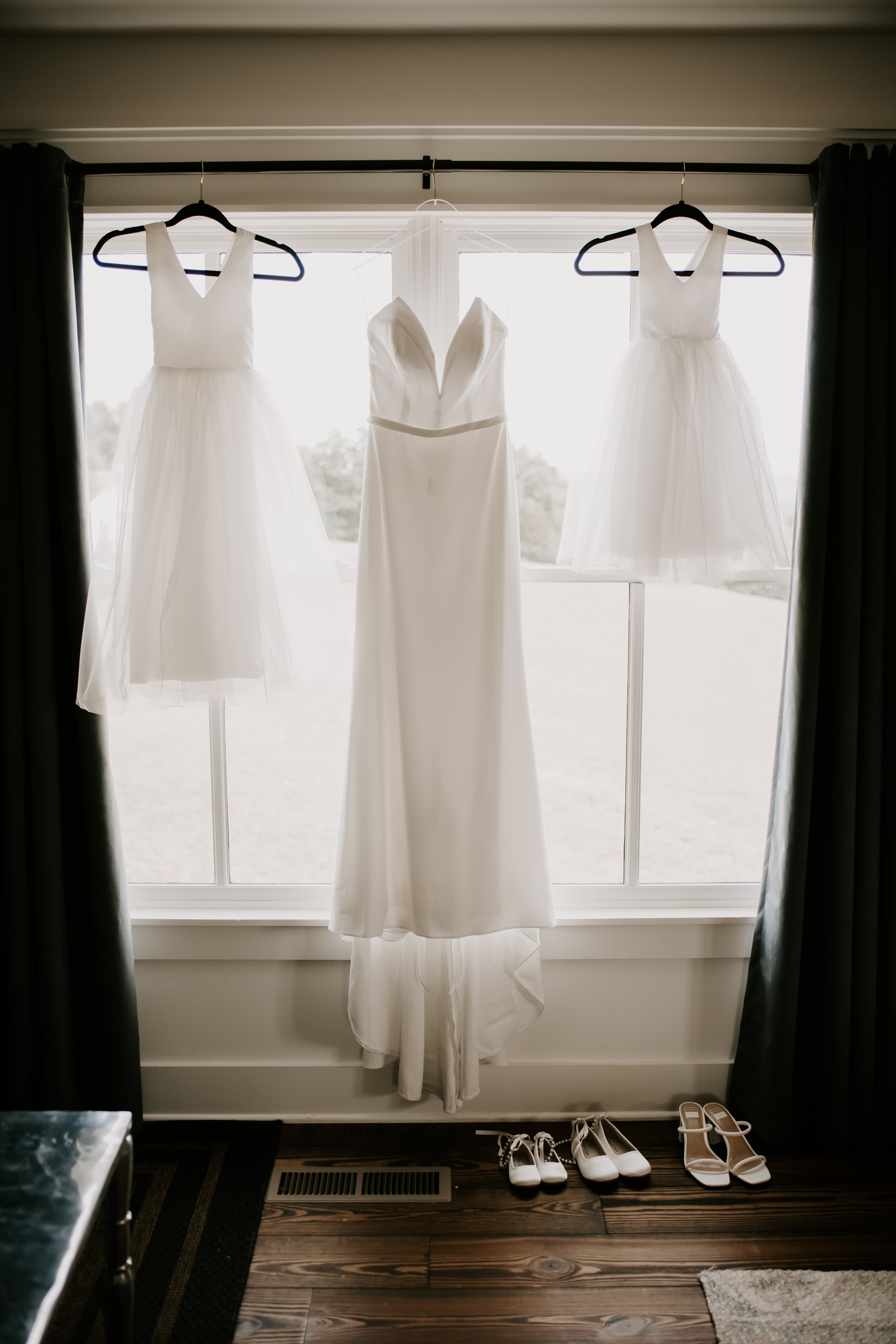 Three white dresses and matching shoes hang by a window.