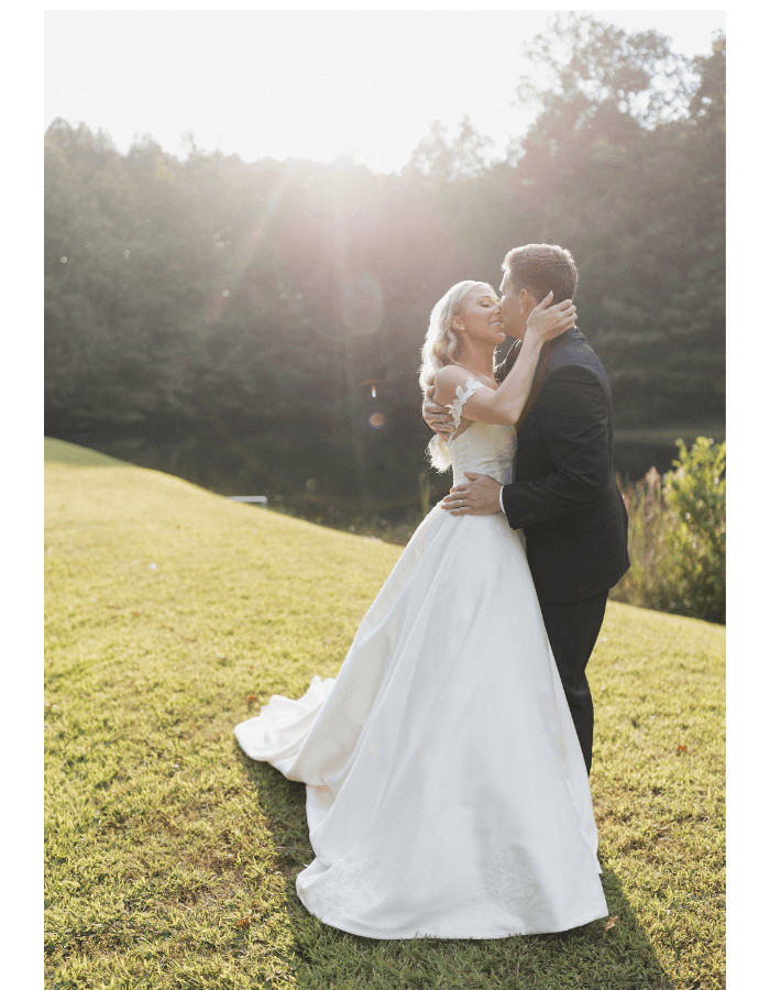 A couple in wedding attire shares a kiss in a sunlit outdoor setting.