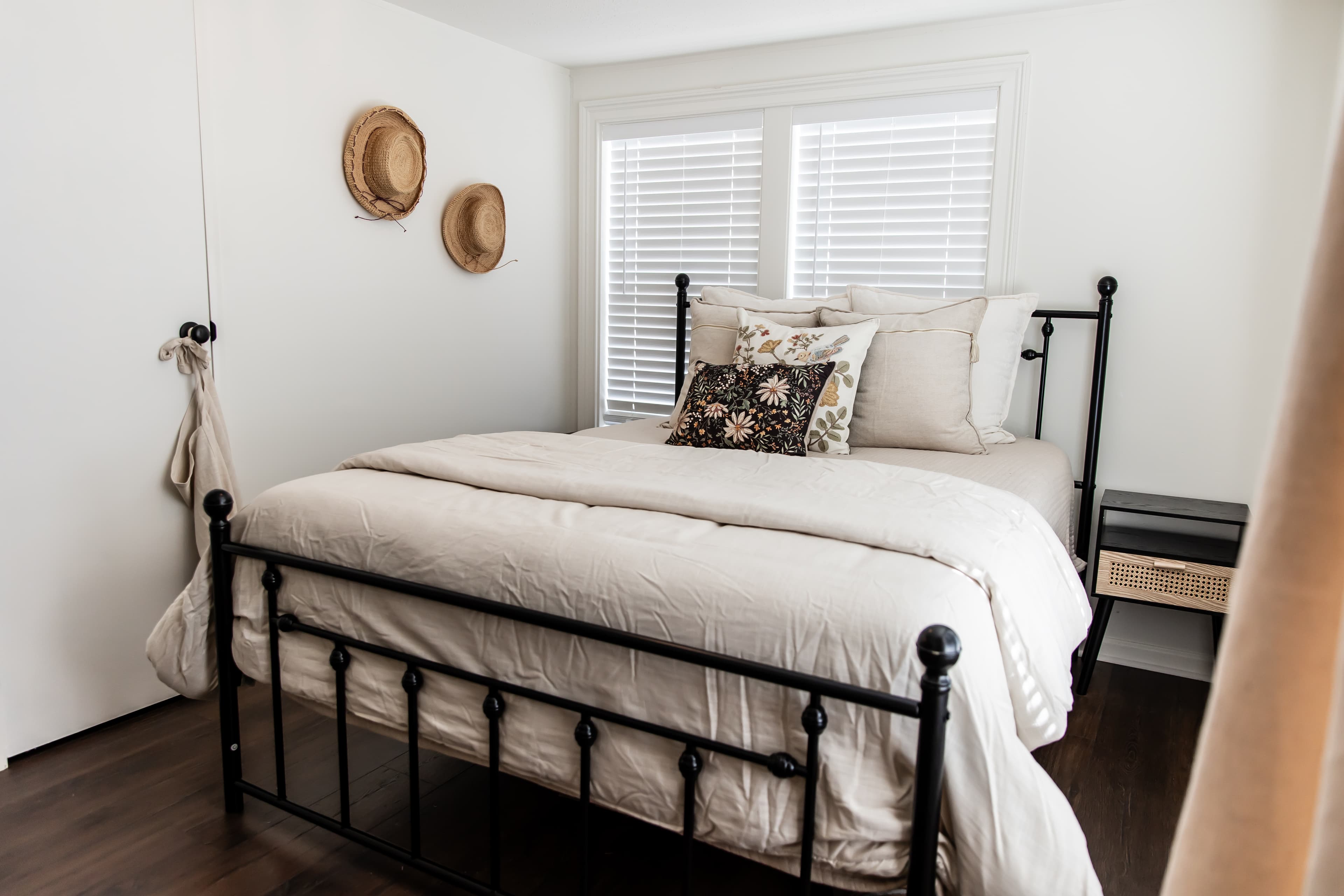 A cozy bedroom featuring a black metal bed with neutral bedding, decorative pillows, and straw hats on the wall.