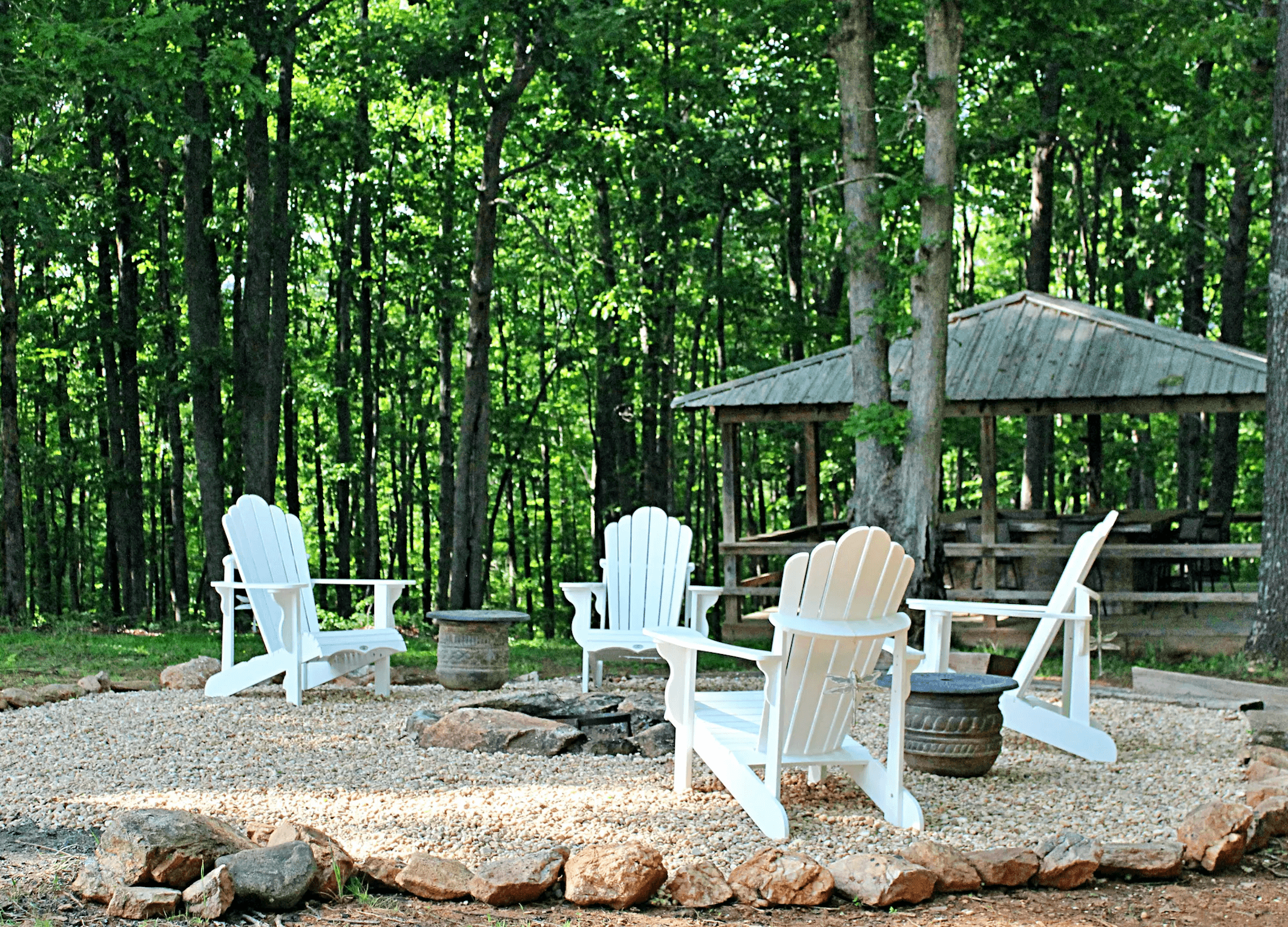 A gravel seating area with white Adirondack chairs surrounding a fire pit in a forested setting.