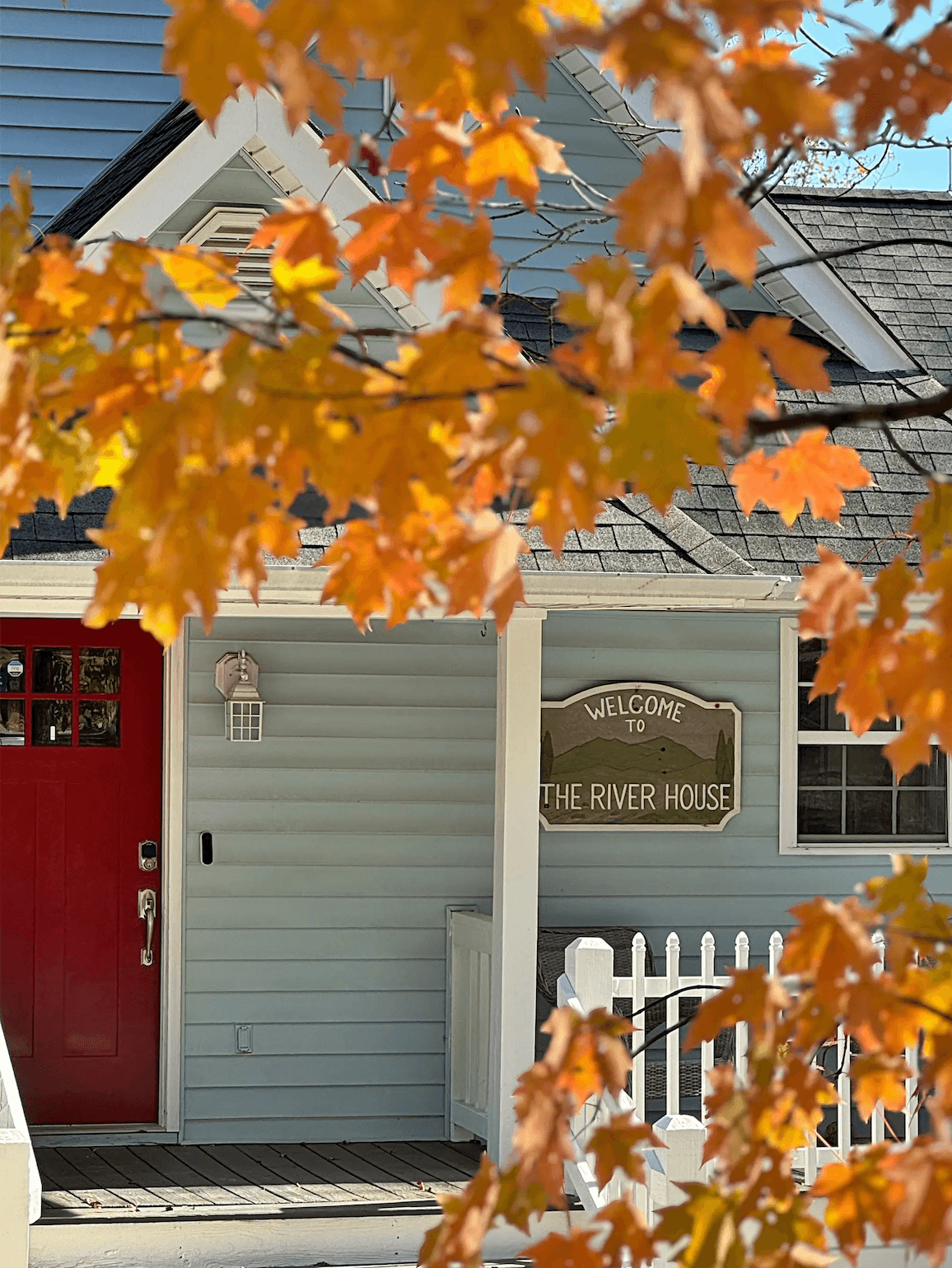 A light blue house with a red door and a sign that reads "Welcome to The River House," framed by autumn leaves.