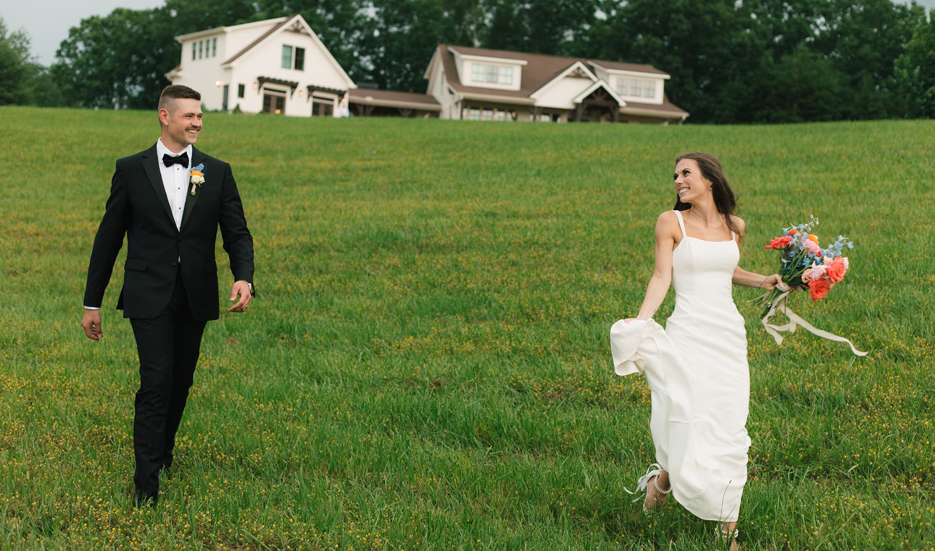 A couple joyfully walks hand in hand in a green field, with a rustic house in the background.