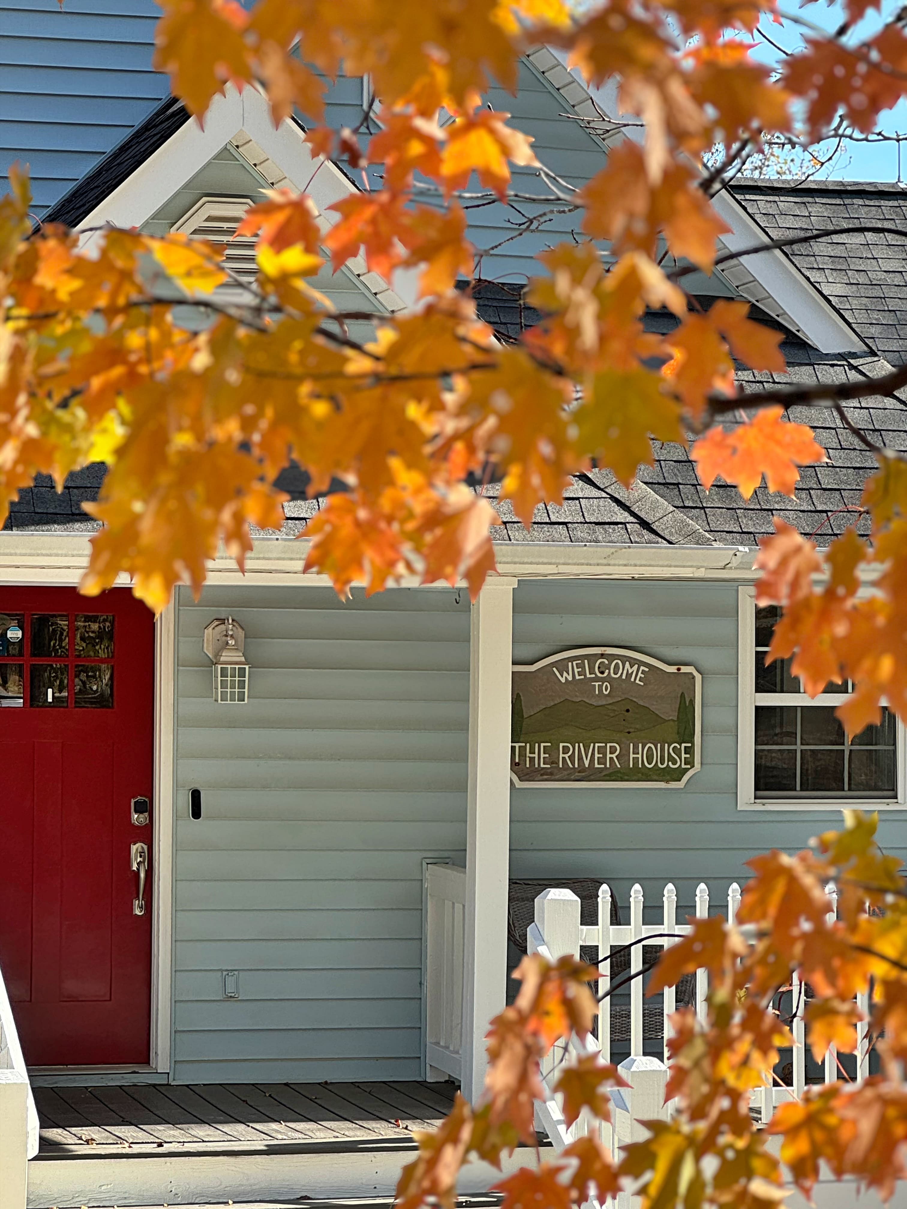 A cozy house with a red door and a "Welcome to The River House" sign, framed by autumn leaves.
