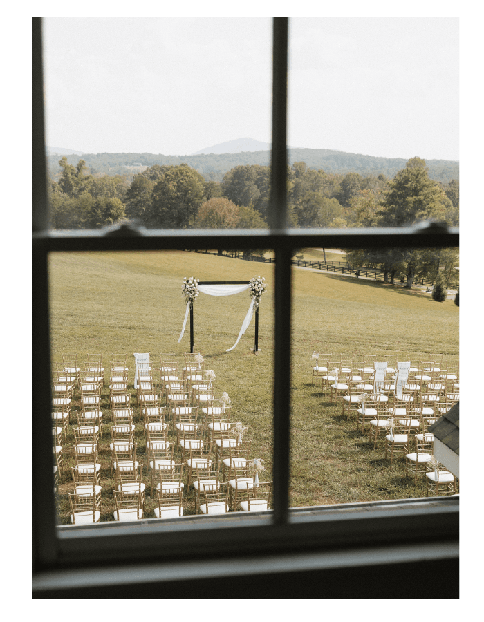 A view through a window of an outdoor wedding ceremony setup with rows of chairs and an arch adorned with flowers.