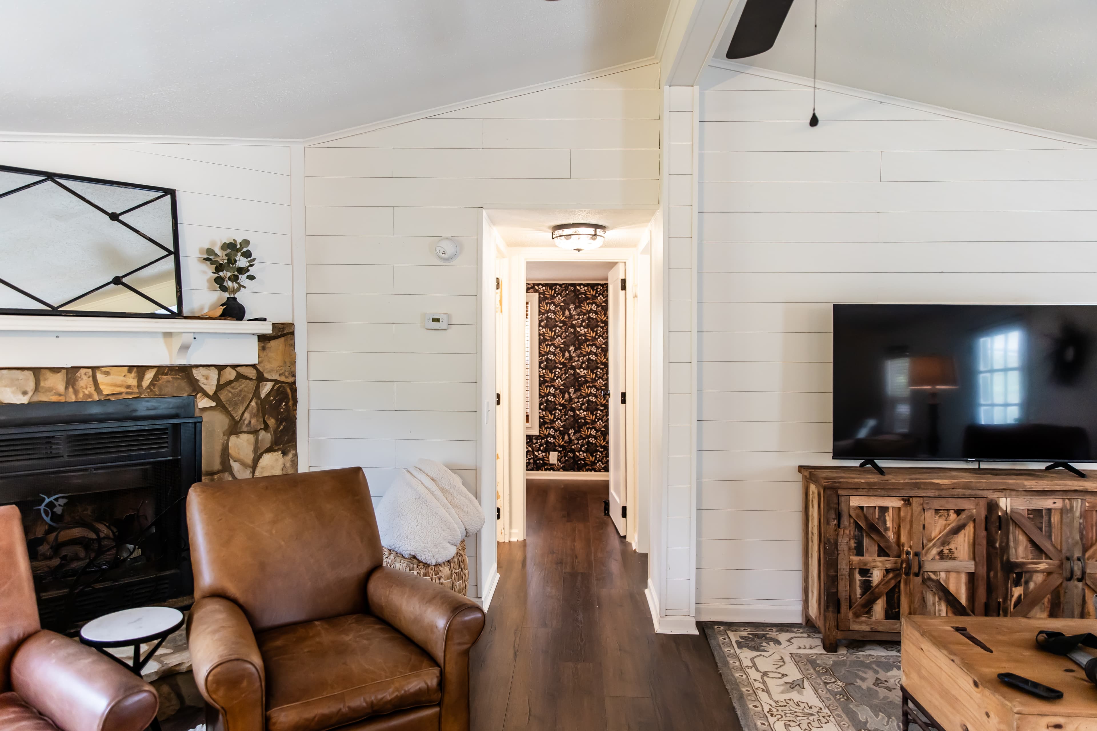 Cozy living room featuring leather armchairs, a stone fireplace, and a large TV, with a view of a hallway.