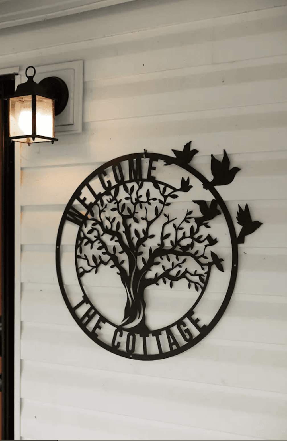 A decorative metal sign featuring a tree and birds with the words "Welcome to the Cottage" on a white wall next to a lantern.