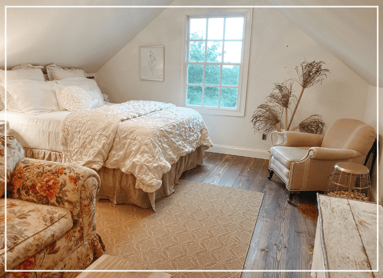 Cozy attic bedroom featuring a large bed, floral armchair, and a window with a view.