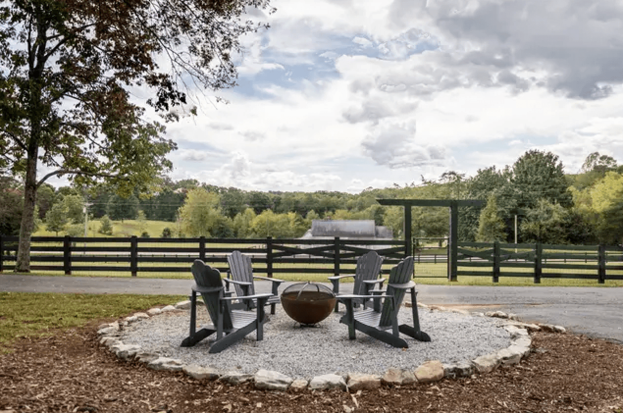 A circular seating area with Adirondack chairs surrounds a fire pit, set against a lush, green landscape.