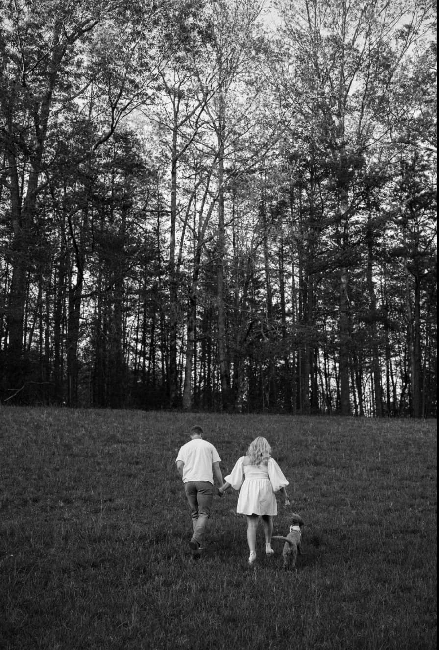 A couple walks hand-in-hand with a small dog through a grassy field toward a forest.