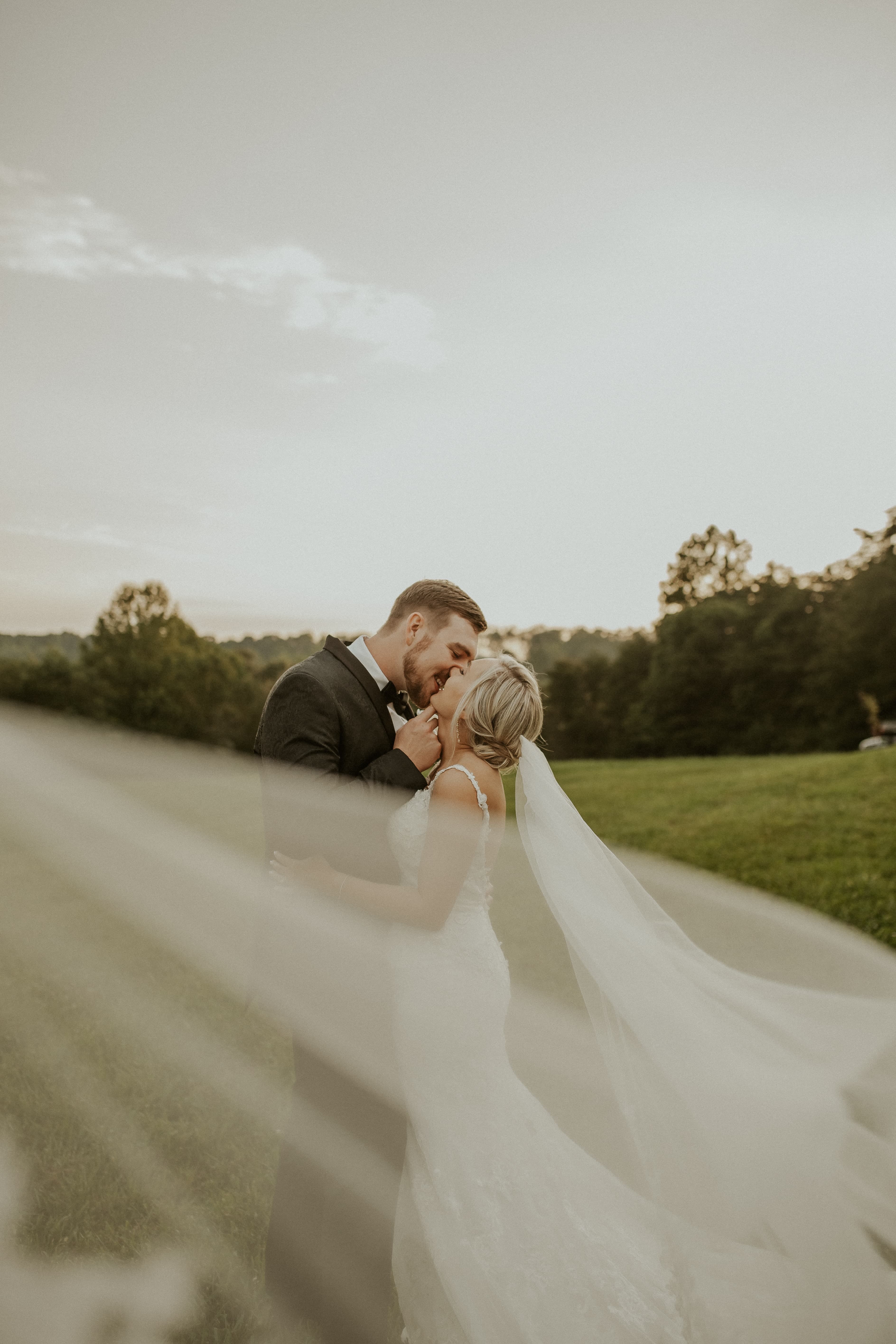A couple shares a romantic kiss in a grassy field, veil flowing in the breeze.