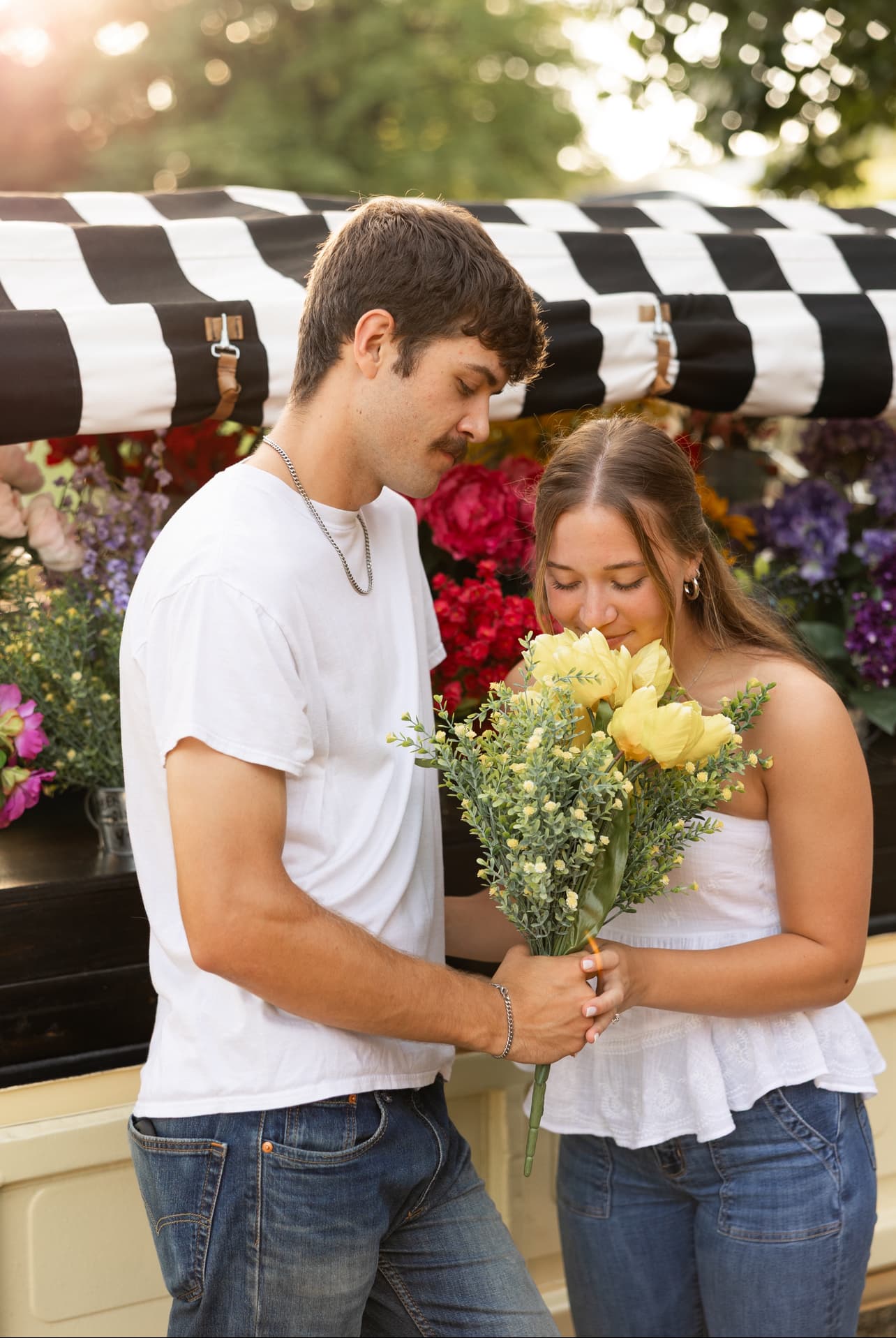 A couple stands close together, with the woman holding a bouquet of yellow flowers while smiling at the man, against a backdrop of colorful flowers.