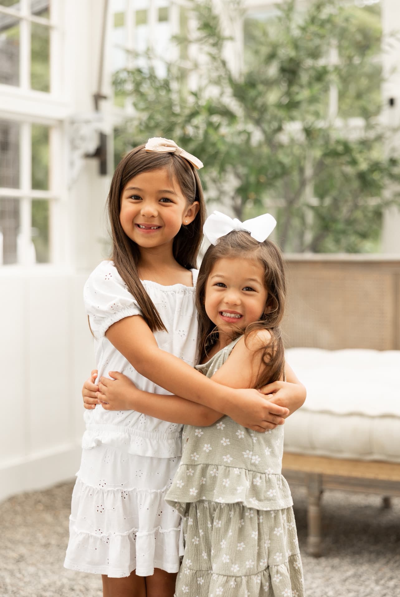 Two young girls embrace each other, both smiling, in a bright indoor setting.