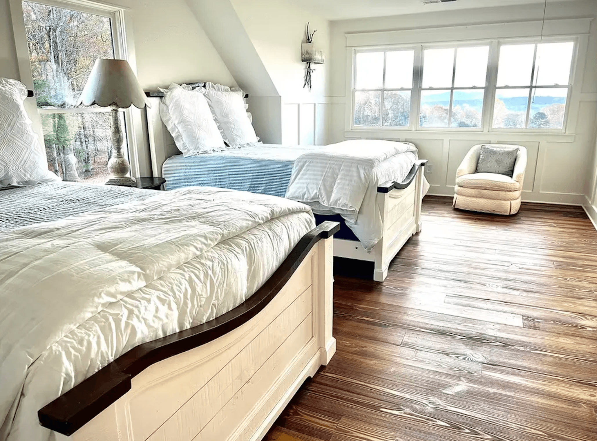 A bright, cozy bedroom features two beds with white linens, a lamp, and a comfortable chair by a window overlooking a scenic view.