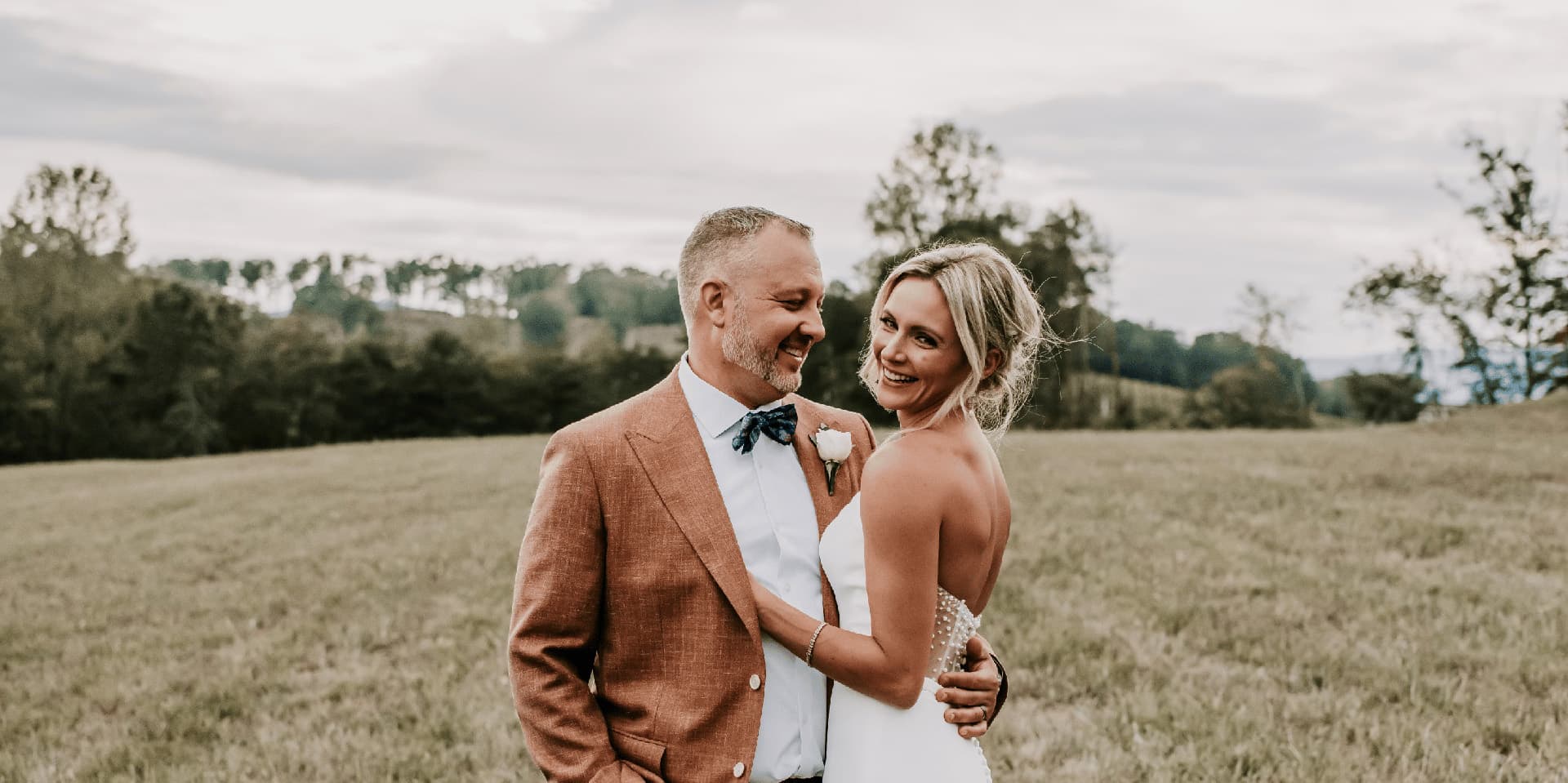 A smiling couple embraces in a grassy field, surrounded by trees and a cloudy sky.