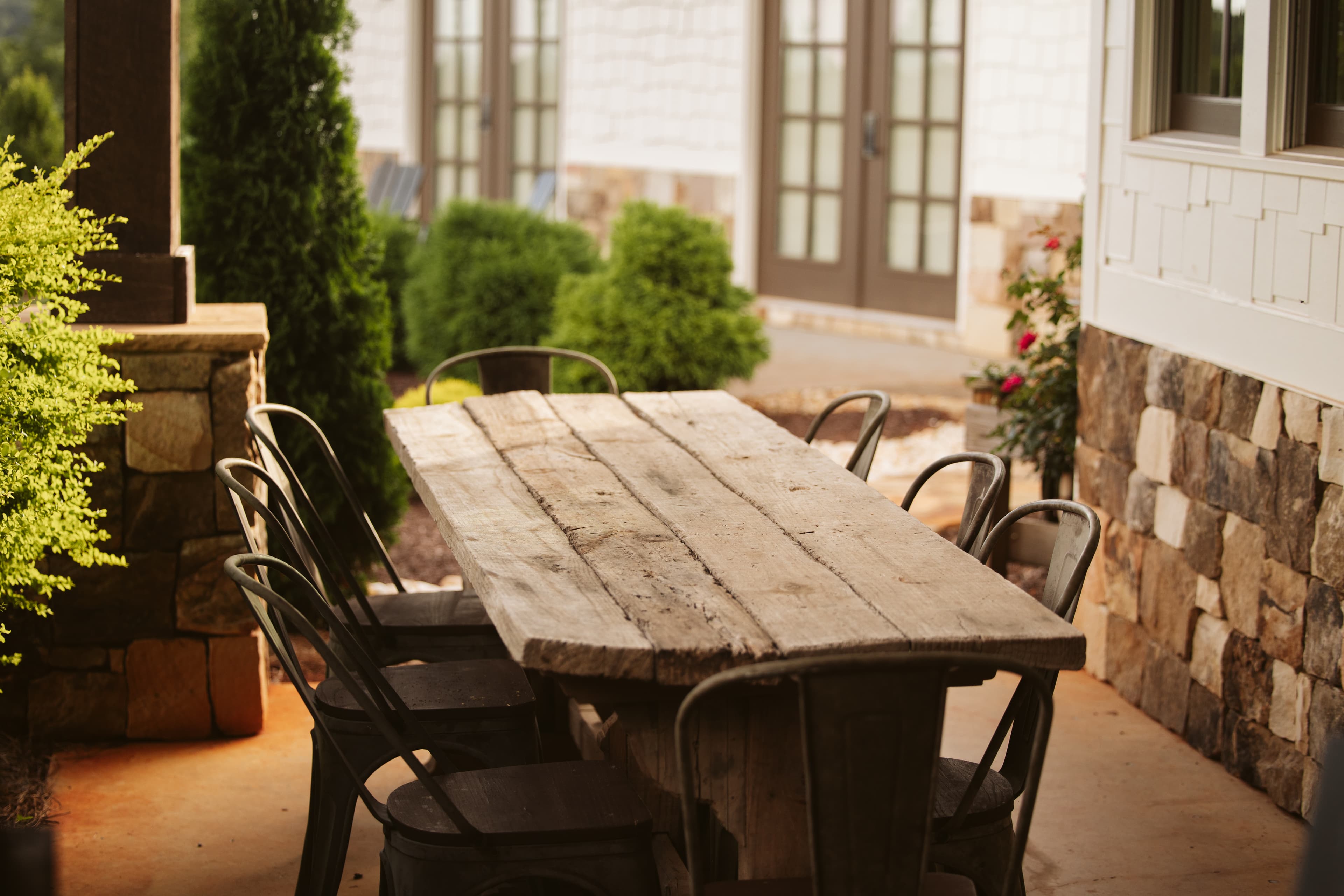 A rustic wooden table with metal chairs is set on a patio surrounded by greenery.