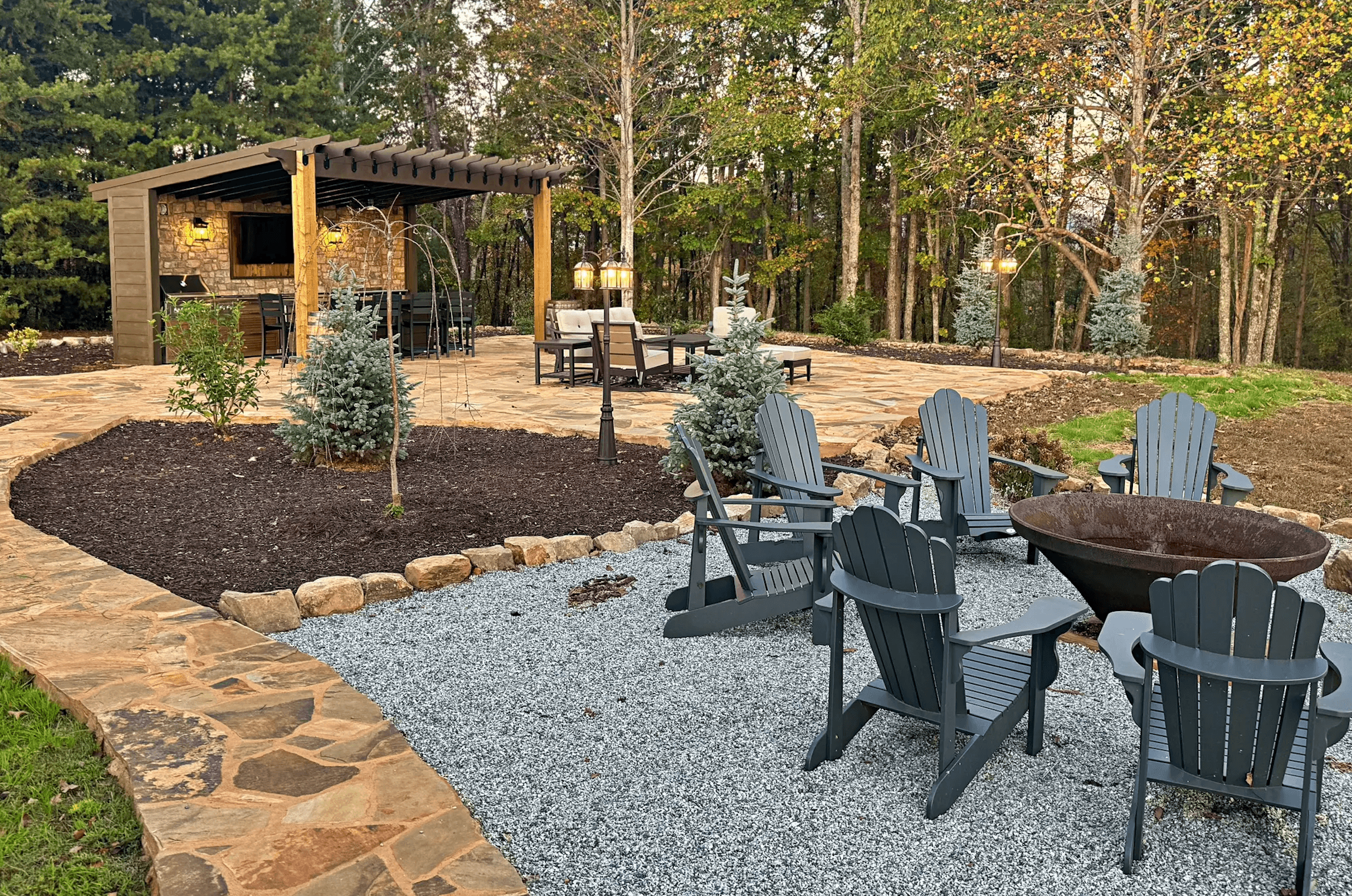 A cozy outdoor space featuring a fire pit surrounded by Adirondack chairs, stone pathways, and a shaded seating area.