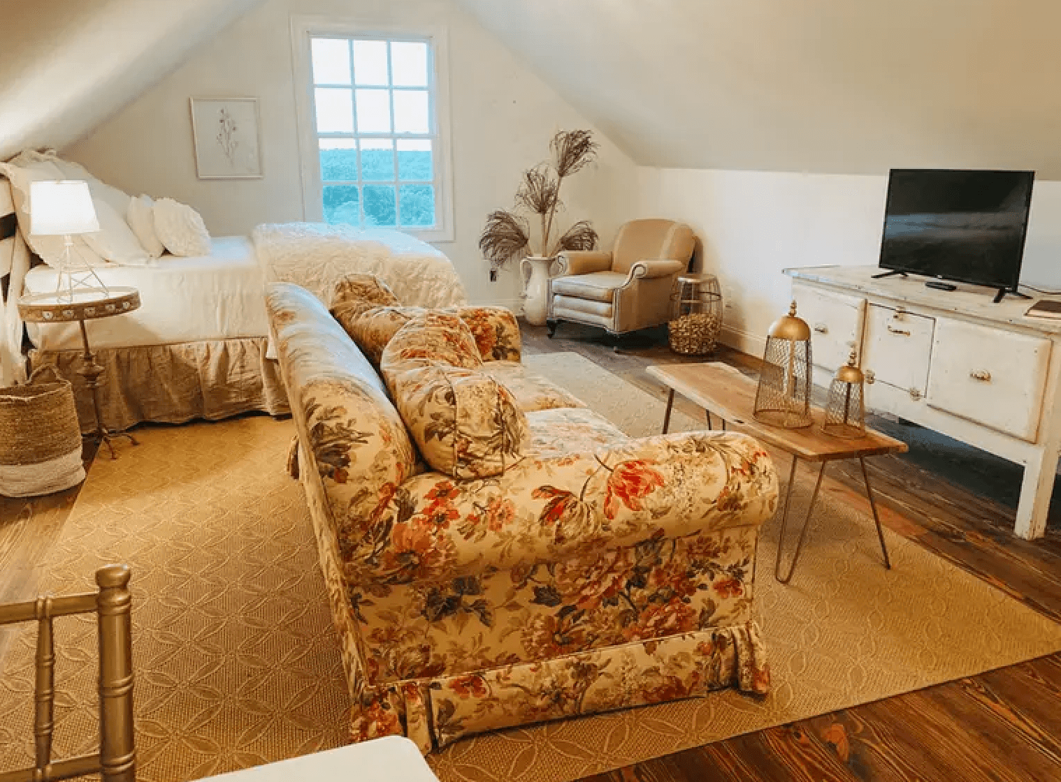 Cozy attic room featuring a floral sofa, a bed, a TV, and natural light from a large window.
