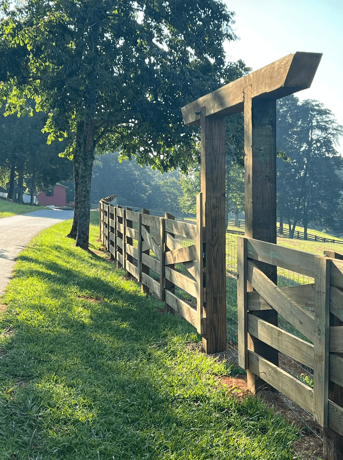 A wooden fence lines a path next to grassy areas and trees under a clear sky.
