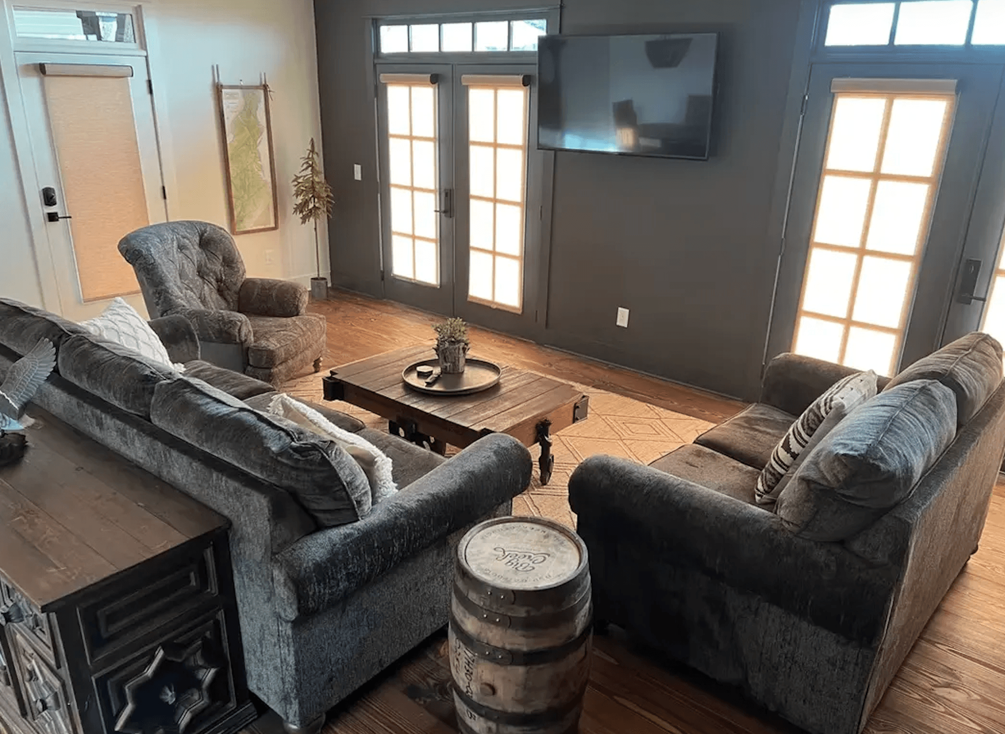 A cozy living room featuring two sofas, a recliner, a round coffee table with a plant, a wooden barrel, and a wall-mounted TV near large windows.
