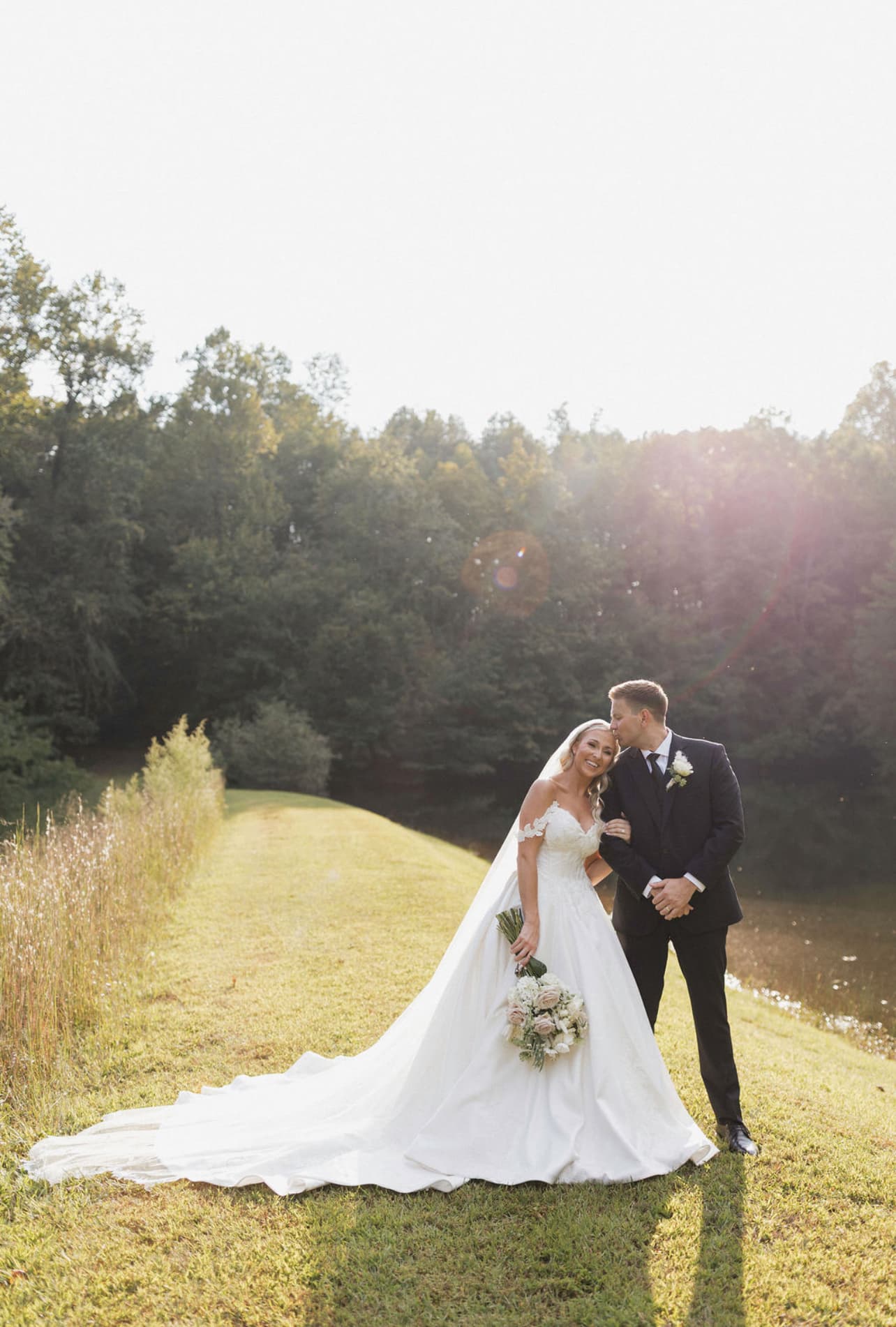 A smiling bride and groom embrace on a grassy path beside a serene pond, illuminated by soft sunlight.
